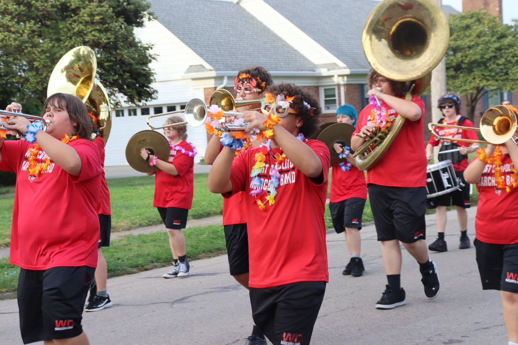 A marching band on the street in front of a house. Members wear red shirts and black shorts and are playing instruments..