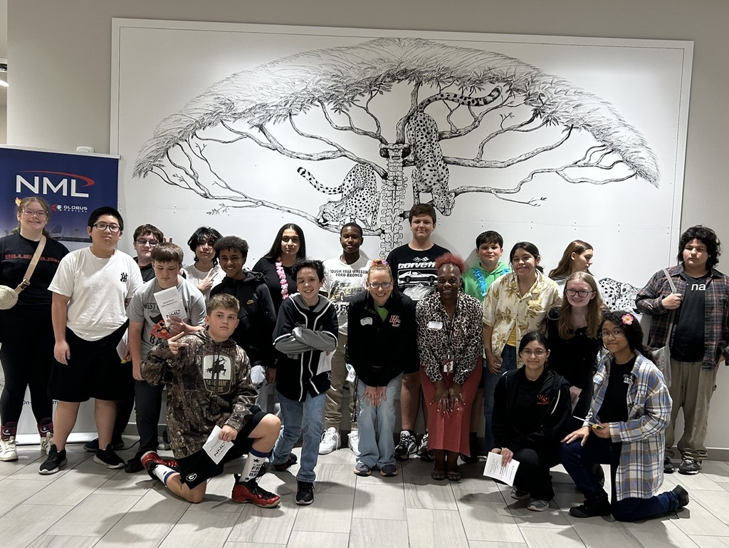 Group of people in front of a white wall with a black drawing of a tree with cheetahs in the branches. A sign for NML is visible in the background.