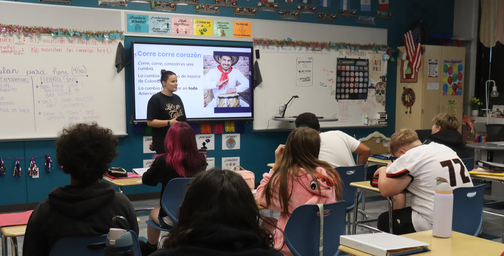Teacher in front of a classroom of students sitting at desks. Whiteboards and signs in the background have Spanish text. A picture on a whiteboard is a smiling man drumming wearing a white shirt with a red scarf.