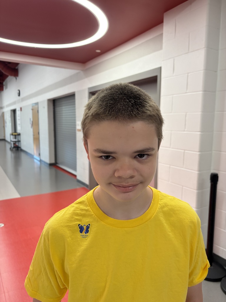 A boy in a yellow t-shirt stands in a hallway with white walls and red flooring.