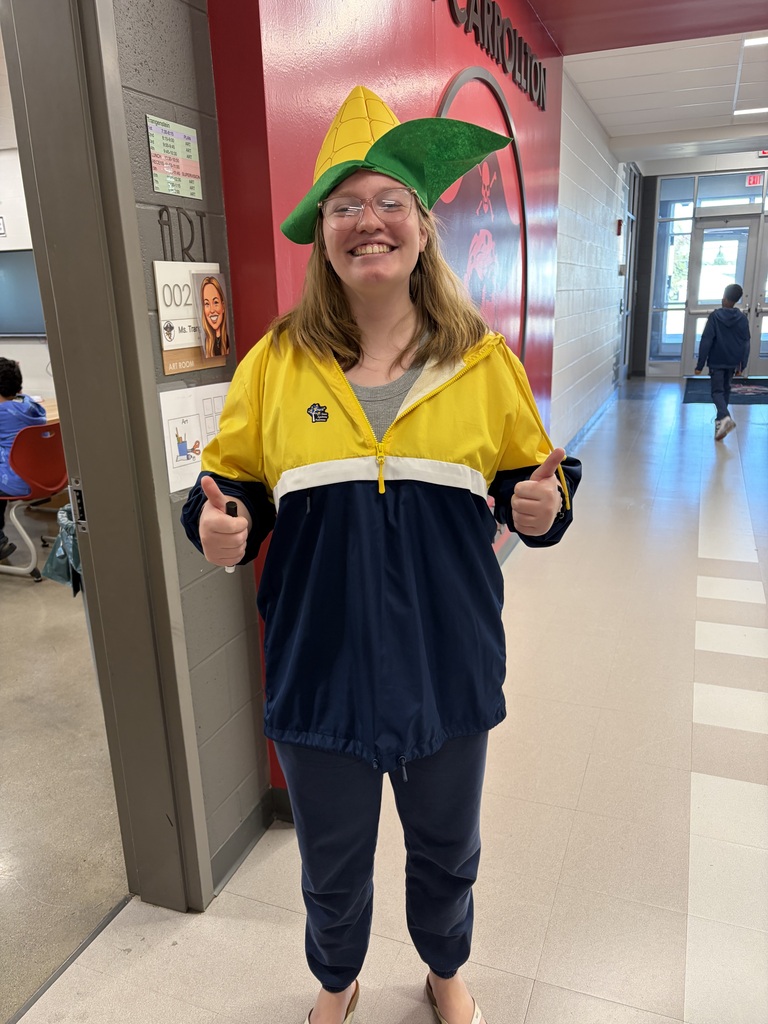 A woman in a yellow and blue jacket, yellow and green hat, and glasses poses with thumbs up in a hallway.