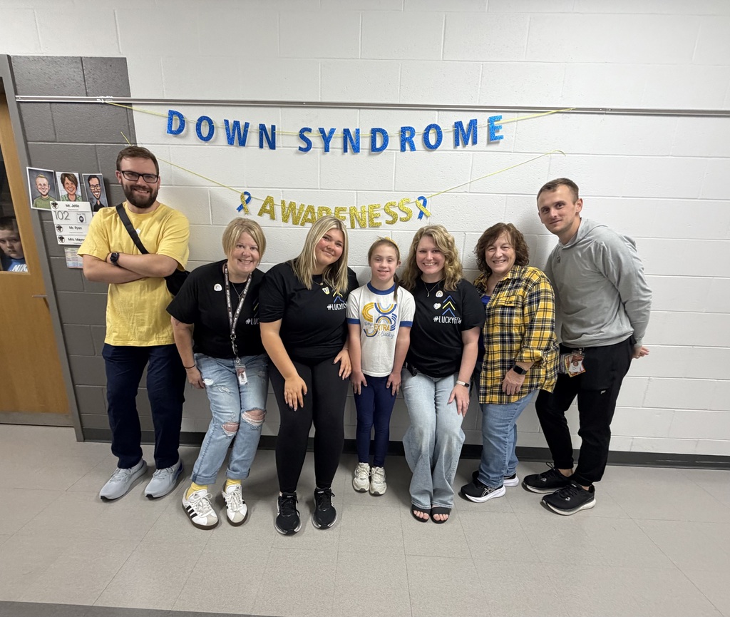 A group of adults with one child standing in the middle stand in front of a wall with Down Syndrome awareness banners.