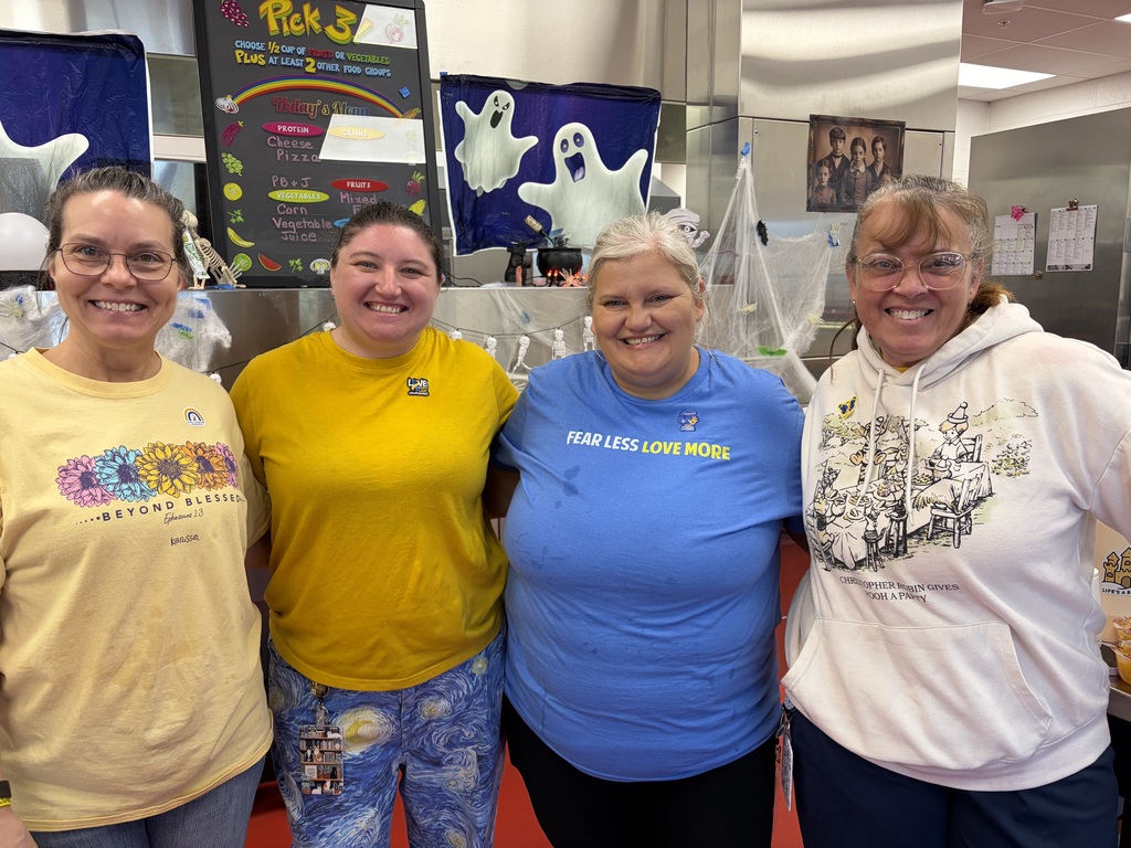 Four women standing together, two wearing yellow t-shirts, one wearing a blue t-shirt, and one wearing a white hoodie, posing together and smiling. Images of a ghost and a menu board are in the background.