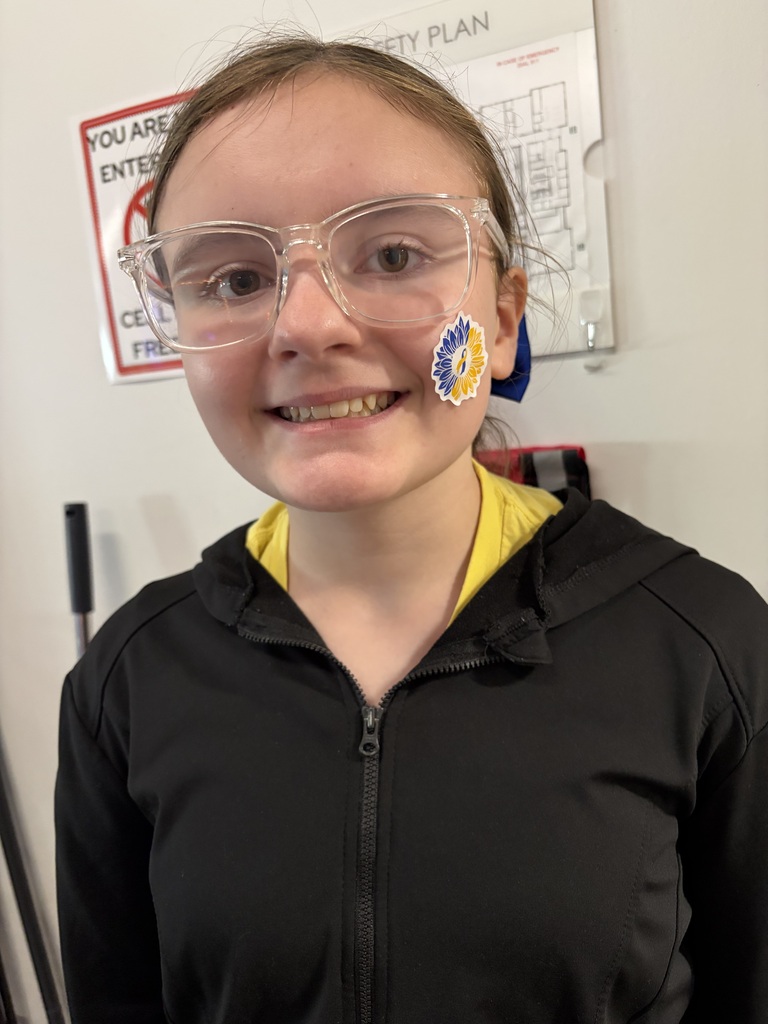 A young girl wearing clear glasses and a yellow and blue sticker on her cheek, wearing a yellow shirt and black jacket, standing in a room with a white wall and a sign.