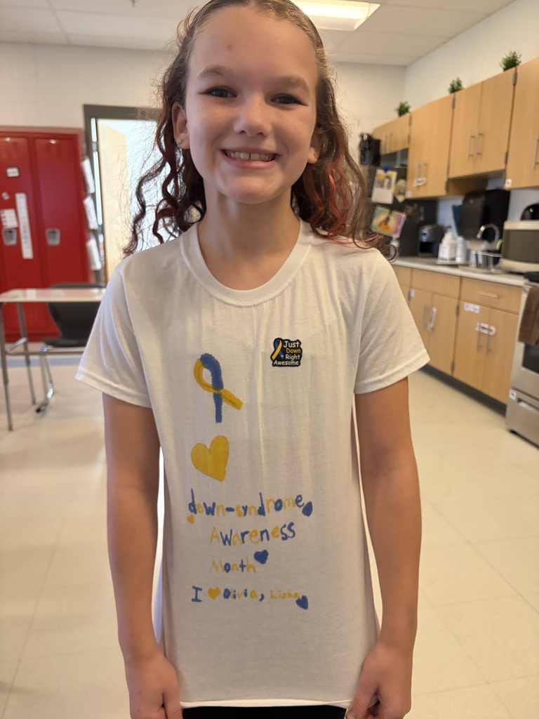 A girl stands in a classroom with a kitchen and cabinets, wearing a white T-shirt with a yellow ribbon and a blue heart, smiling.