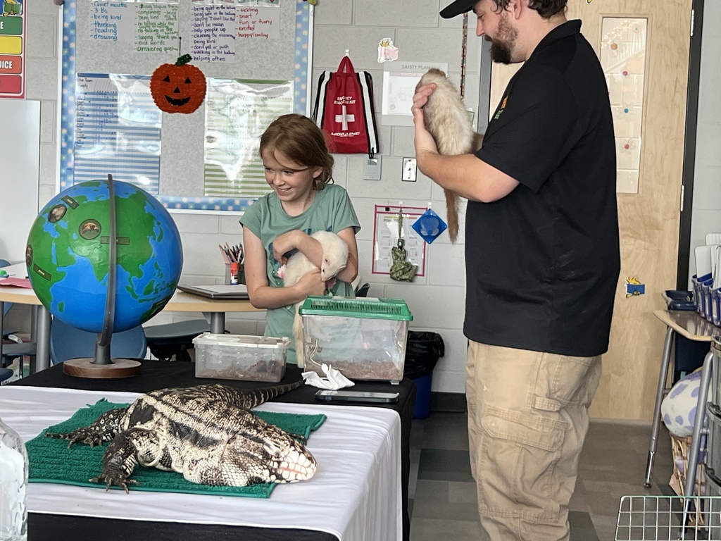 Two people hold ferrets in a classroom. Display includes a globe and table with a lizard.