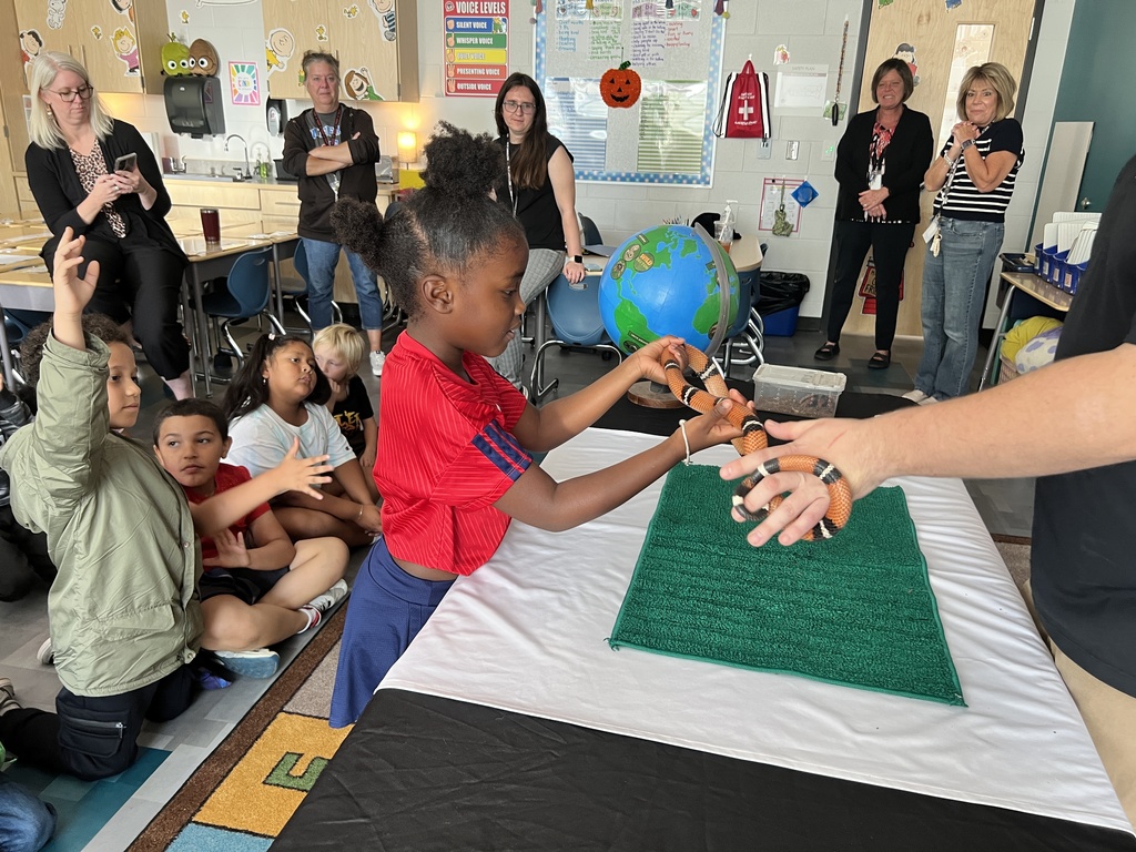 Children sit around a table with a globe, a child holds a snake while an adult assists.