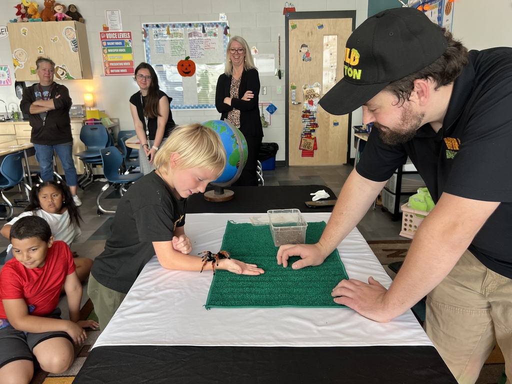 A man helps a child touch a spider on a table in a classroom. Other children and adults are in the background.