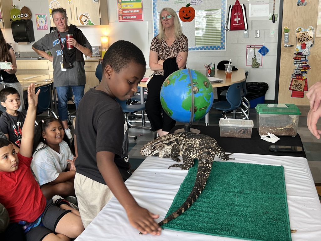 Children and adults in a classroom setting. A boy is touching a lizard. A globe is on the table.