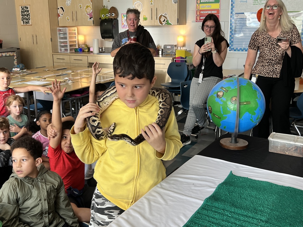 A young boy holds a snake in a classroom. Children sit on the floor behind him, while teachers stand nearby.
