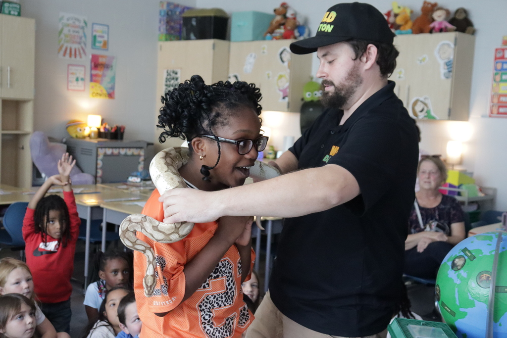 A man puts a snake around a girl's neck. The girl is wearing glasses and an orange shirt. The man is wearing a dark polo. Children sitting on the floor are in the background.