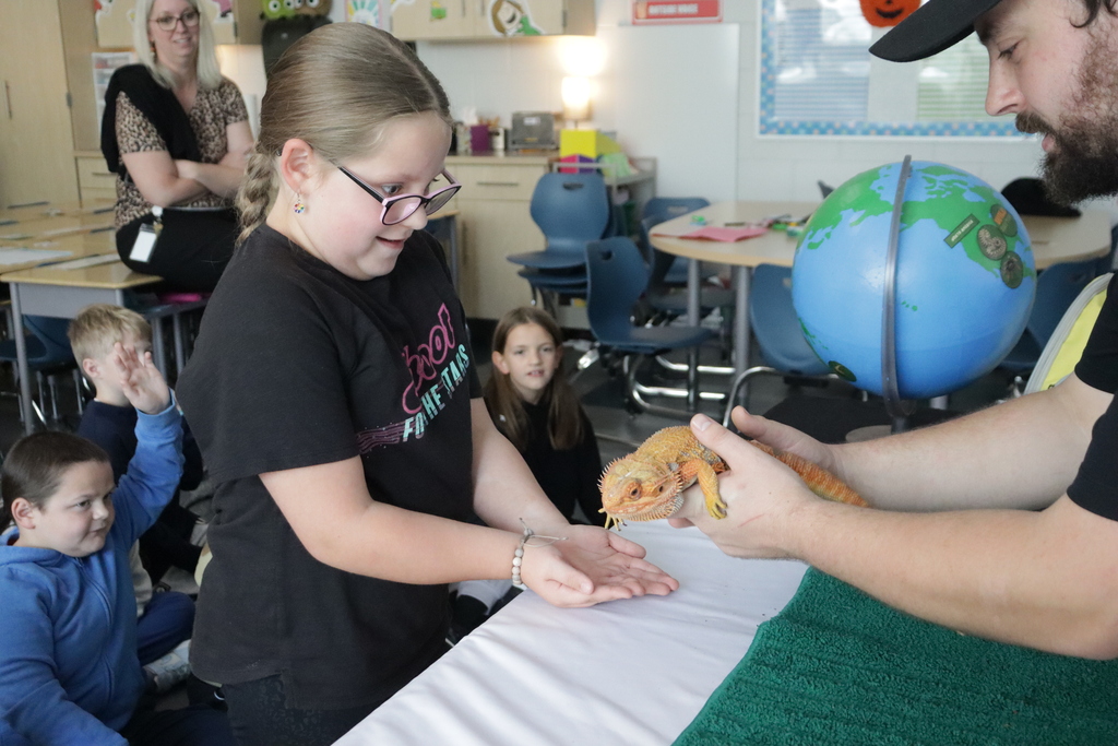 A girl wear in a black shirt wearing glasses holds her hand out to a man with a beard who is handing her a lizard. The globe sits on a table.