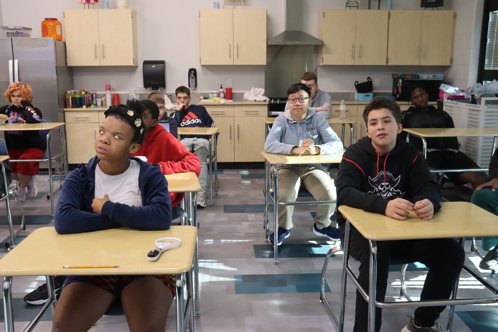 Students in a classroom sit at desks, one has a magnifying glass on the desk.