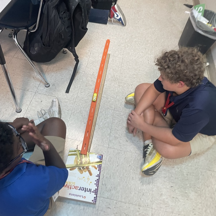 groups of students work on the floor beside an orange Hot Wheels track and a ruler. One group also has a laptop open to record their work.