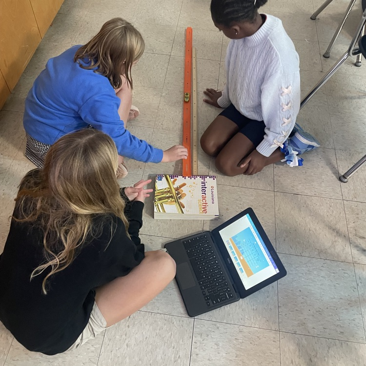 groups of students work on the floor beside an orange Hot Wheels track and a ruler. One group also has a laptop open to record their work.