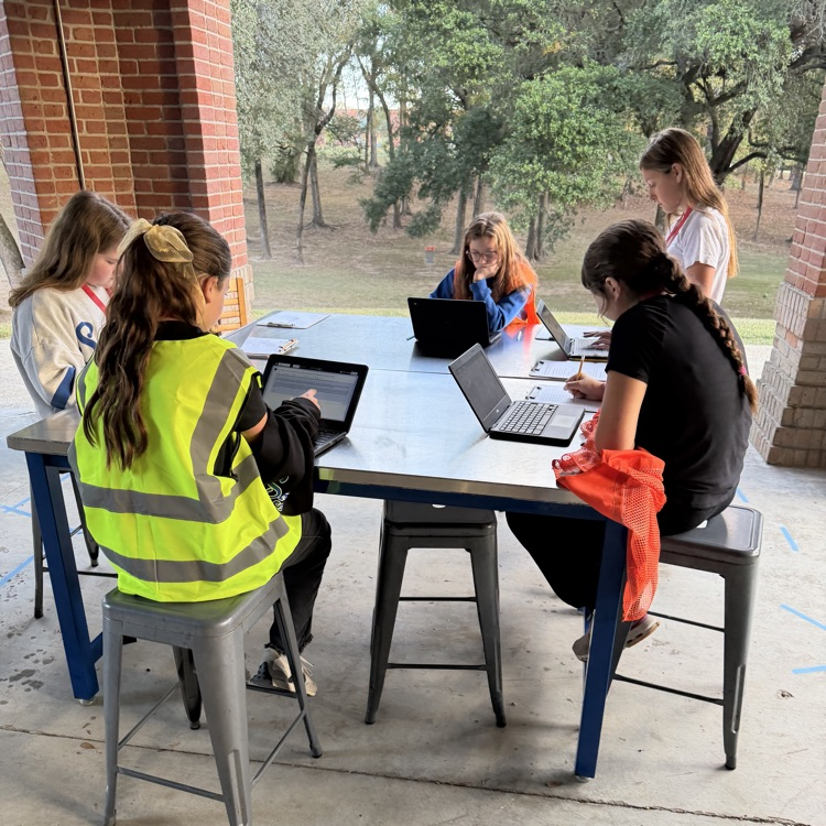 A group of students are seated around a table as they work on computer computers in an outdoor classroom