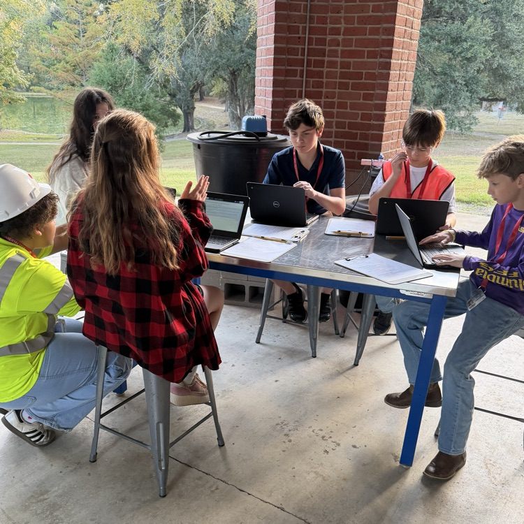 A group of students are seated around a table as they work on computer computers in an outdoor classroom