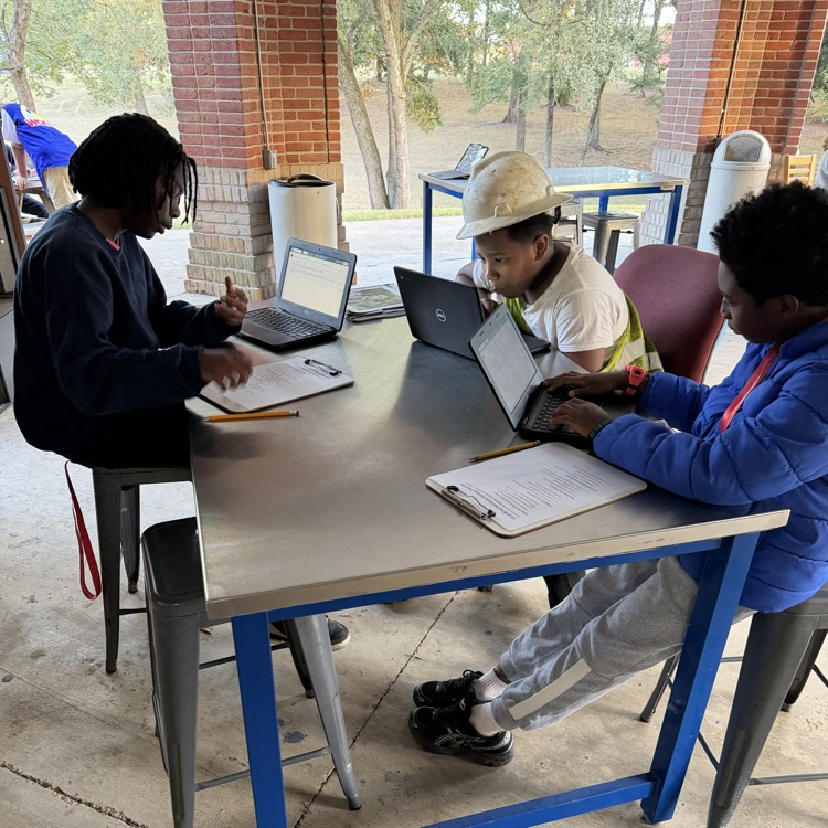 A group of students are seated around a table as they work on computer computers in an outdoor classroom