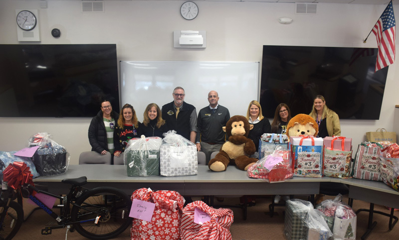 Eight people are standing behind a table filled with wrapped gifts, gift bags, and two large stuffed animals. The mood is festive and cheerful.