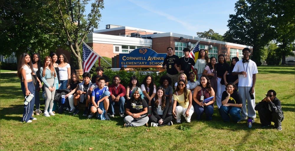 A group of people stands in front of Cornwell Avenue Elementary School with American flags.