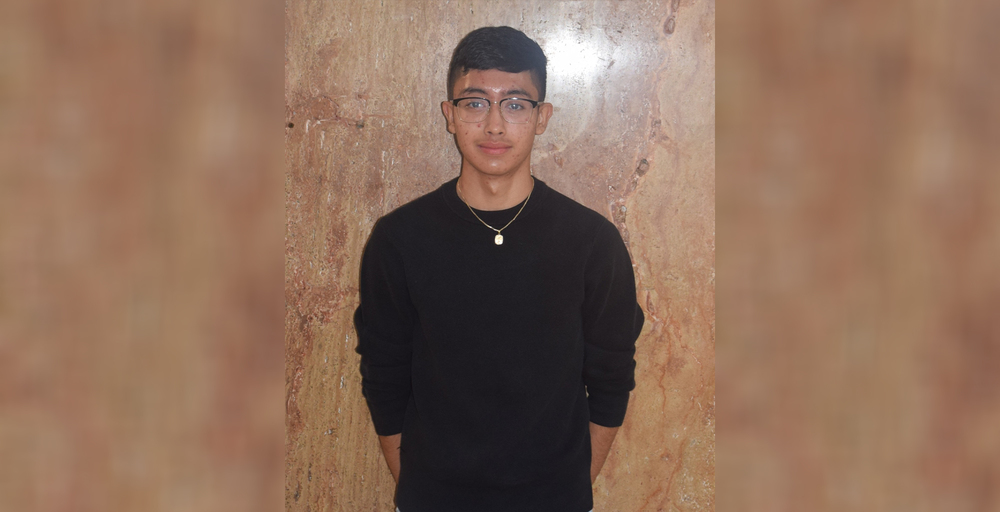 Young man wearing glasses and a black sweater stands against a textured wooden wall, exuding a calm, poised expression; minimalistic and neutral tone.