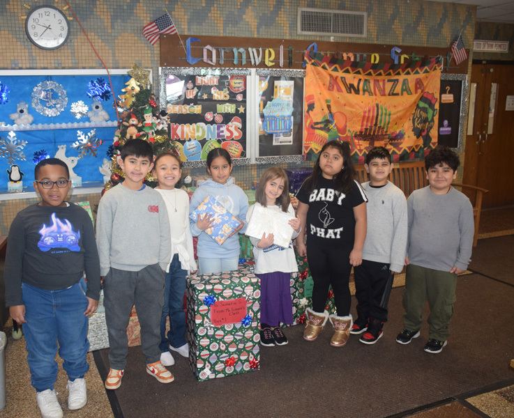 A group of seven young children stand smiling in front of festive holiday decorations, including a blue winter mural, a Kinara banner, and wrapped gifts.