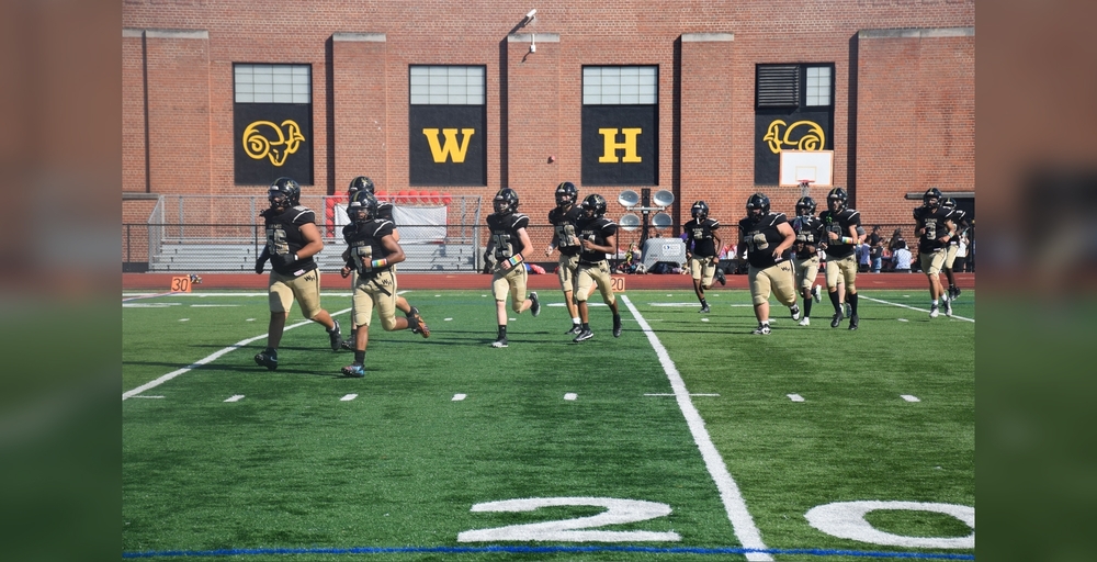 A group of football players running on a field with a building and bleachers in