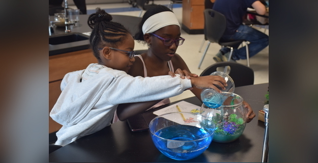 Two girls collaborating in a classroom science experiment, pouring a blue liquid into a fishbowl while surrounded by colorful decorations and a notebook for notes.