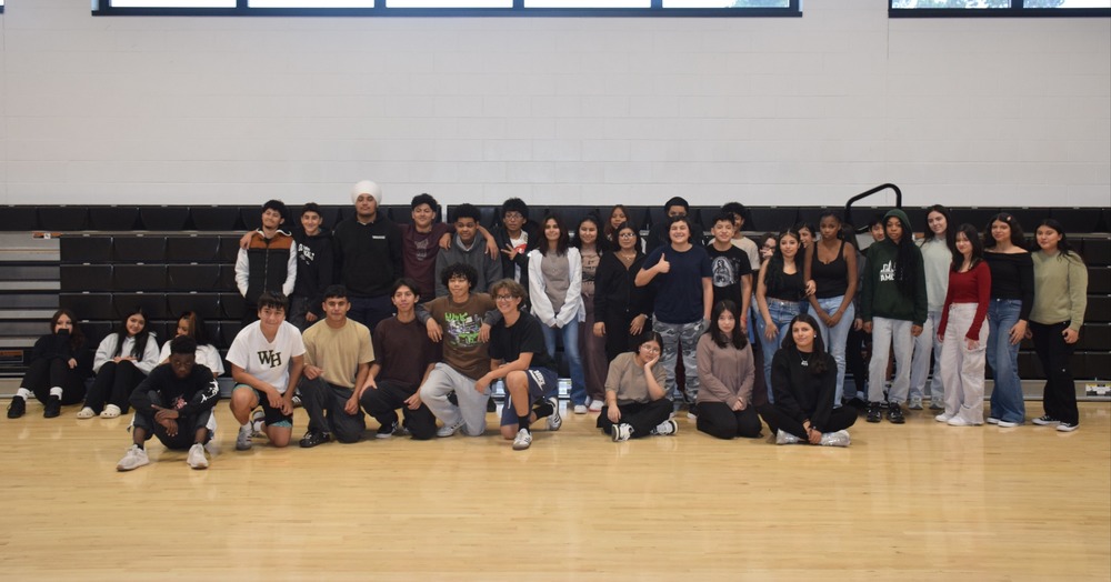 A group of students poses in a gym, with most standing and others sitting on the wooden floor. They look relaxed and cheerful, conveying a sense of unity.