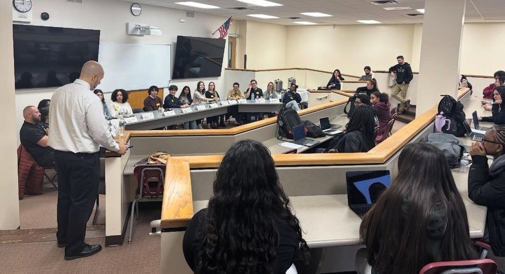 A diverse group of people, seated in a semi-circular classroom, listen attentively to a speaker. Laptops and notebooks are open, creating a focused and collaborative atmosphere.