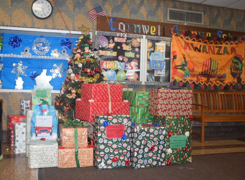 A group of seven young children stand smiling in front of festive holiday decorations, including a blue winter mural, a Kinara banner, and wrapped gifts.