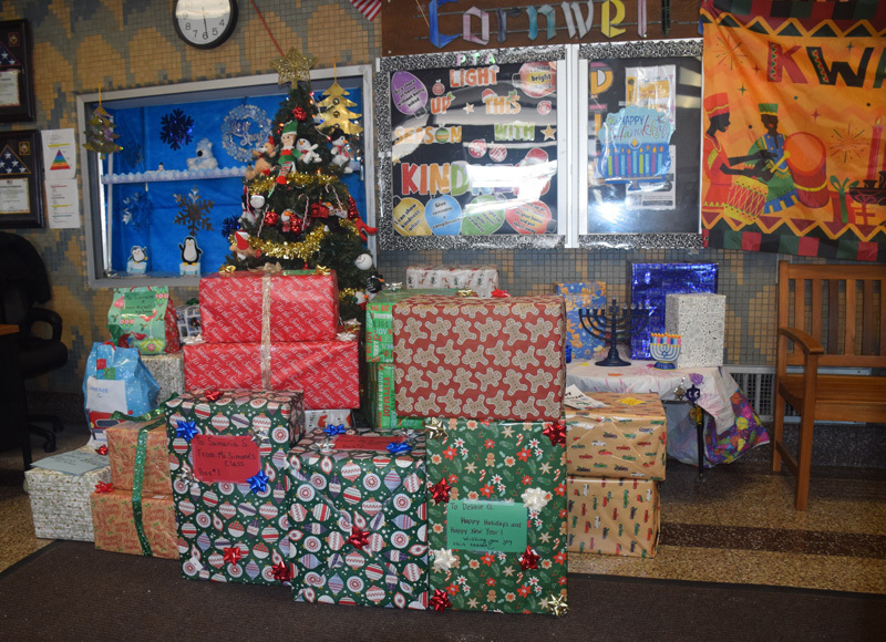 A group of seven young children stand smiling in front of festive holiday decorations, including a blue winter mural, a Kinara banner, and wrapped gifts.