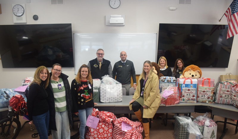 people are standing behind a table filled with wrapped gifts, gift bags, and two large stuffed animals. The mood is festive and cheerful.
