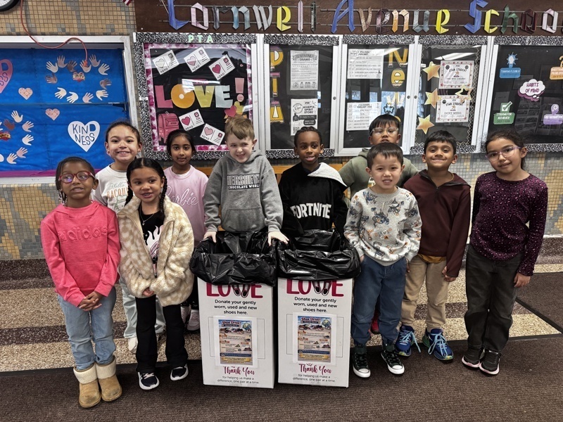 A group of smiling children stands in a school hallway in front of donation boxes