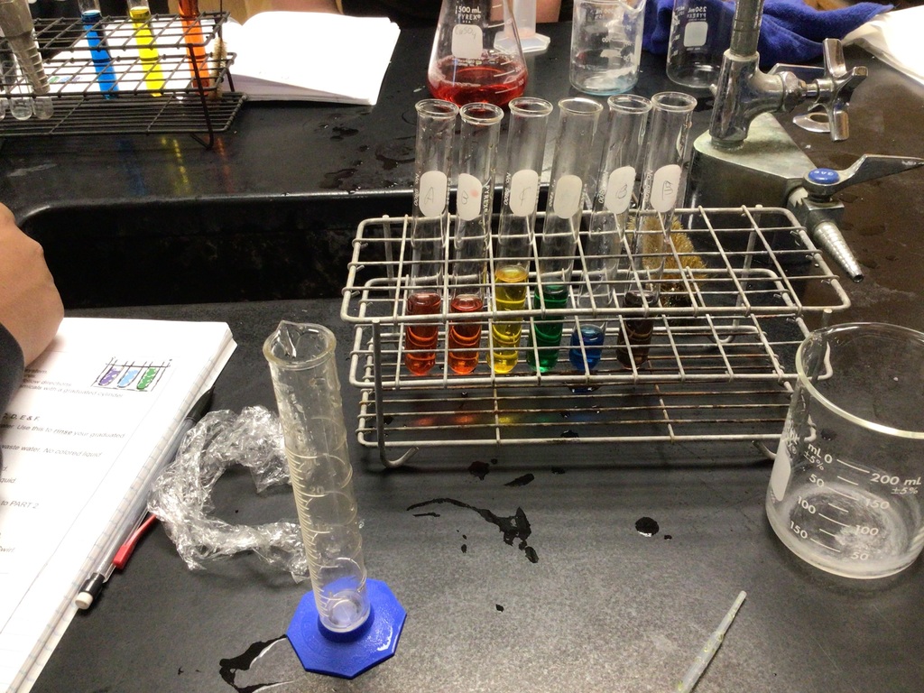 Six test tubes in a rack display liquids in the colors of a rainbow. Other lab equipment is visible on the lab station like beakers, graduated cylinders, pipets, and an erlenmeyer flask containing red liquid.