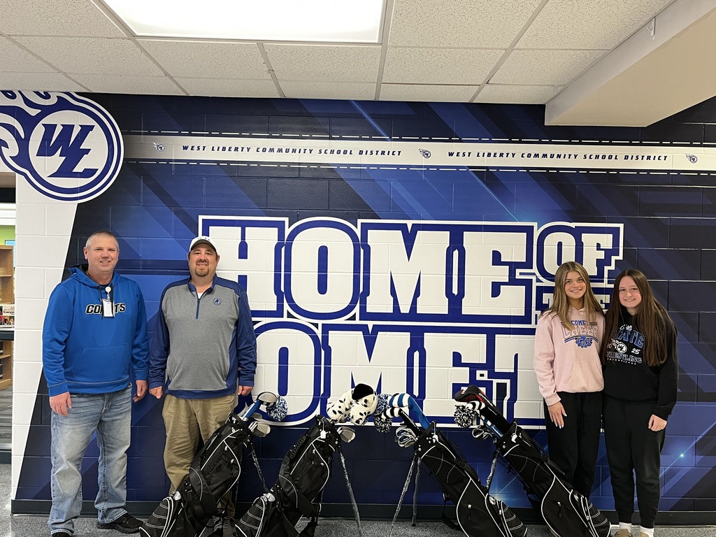 Coach James Laughlin, Clint Brown Iowa Golf Association Director of Marketing and Communication, four complete sets of donated golf clubs and Comet Varsity Girls Golf team members Kamryn Gilmore and Kenley Harned in front of the "Home of the Comets" wall at West Liberty High School