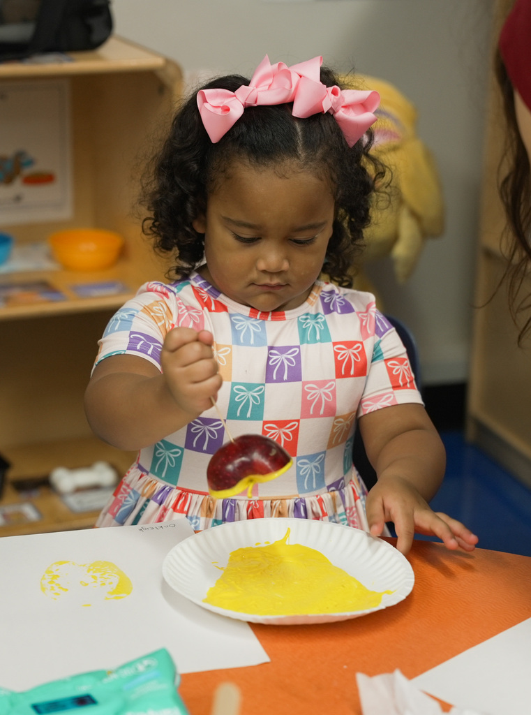 girl with bows in her hair dipping an apple in yellow paint