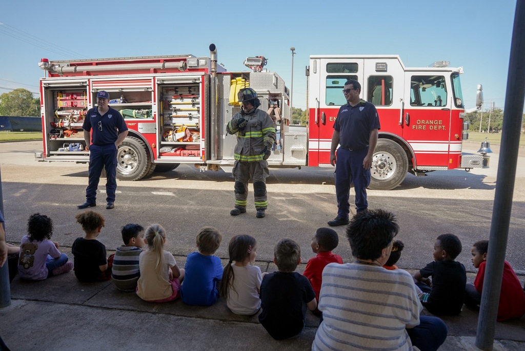 three firefighters in front of a fire truck talking to a group of young children
