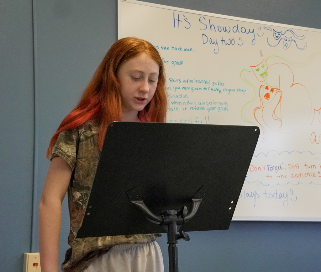 students performing a play, girl with red hair at lectern in front of whiteboard