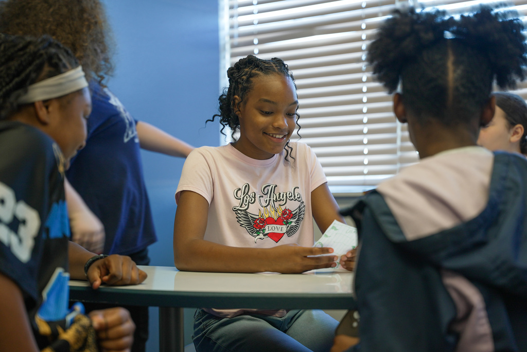 students performing a play, girl sitting at table surrounded by others, reading her script