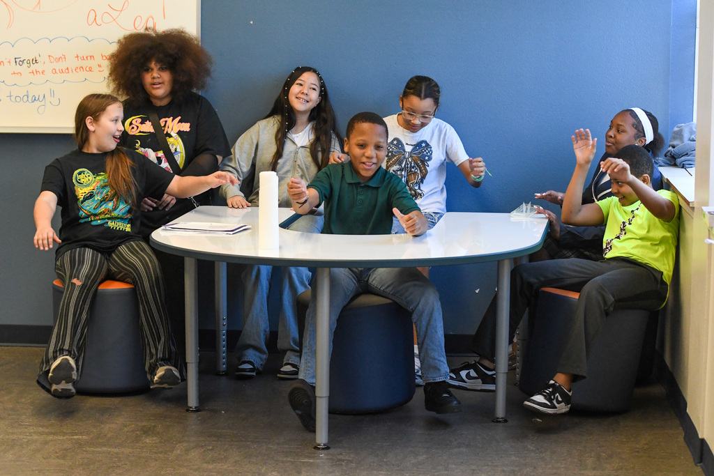 students performing a play, they're seated around a table pretending it's car driving really fast