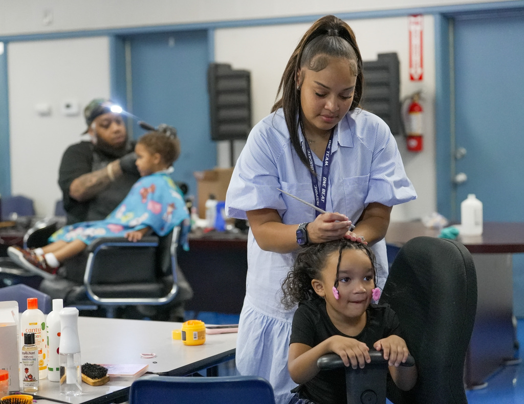 a girl gets her hair worked on in the foreground as a barber works on another boys hair in the back