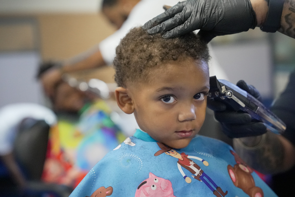 a boy looks directly into the camera as a barber shaves his hair