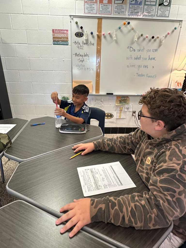 Two students work together during a science lab, observing a balloon inflate as a reaction occurs inside a bottle.