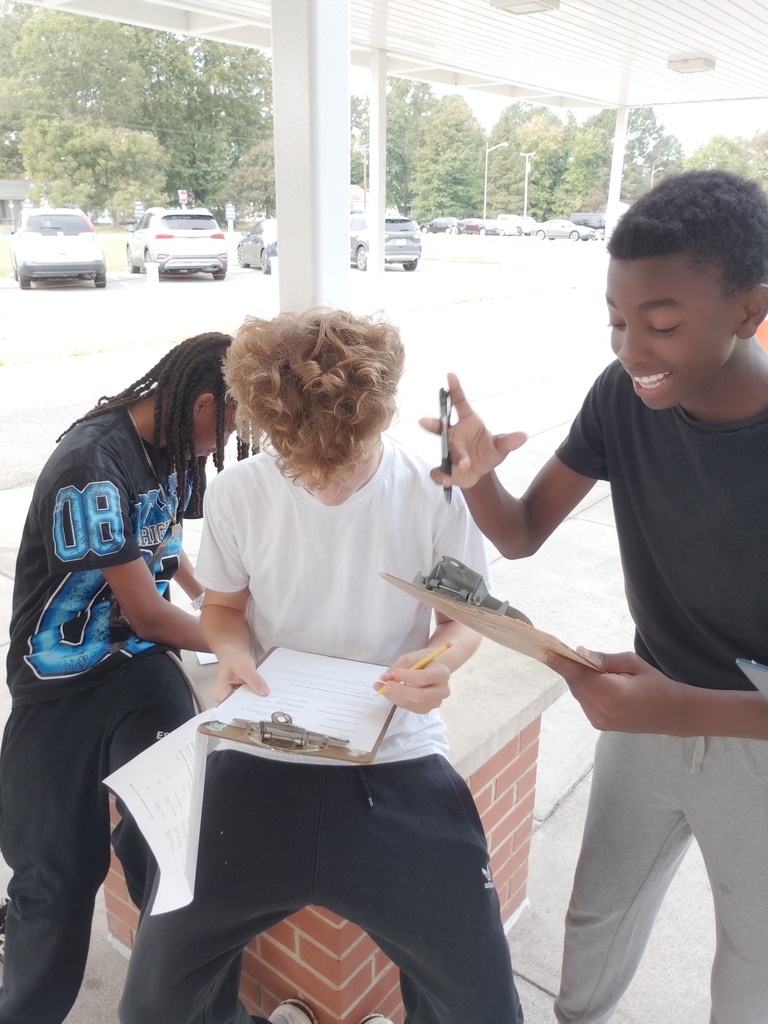 Three students work together outdoors under a covered walkway. One student sits on a low brick ledge filling out a paper on a clipboard with a pencil, while another stands nearby gestures enthusiastically with a pen, and a third student takes notes in the background.