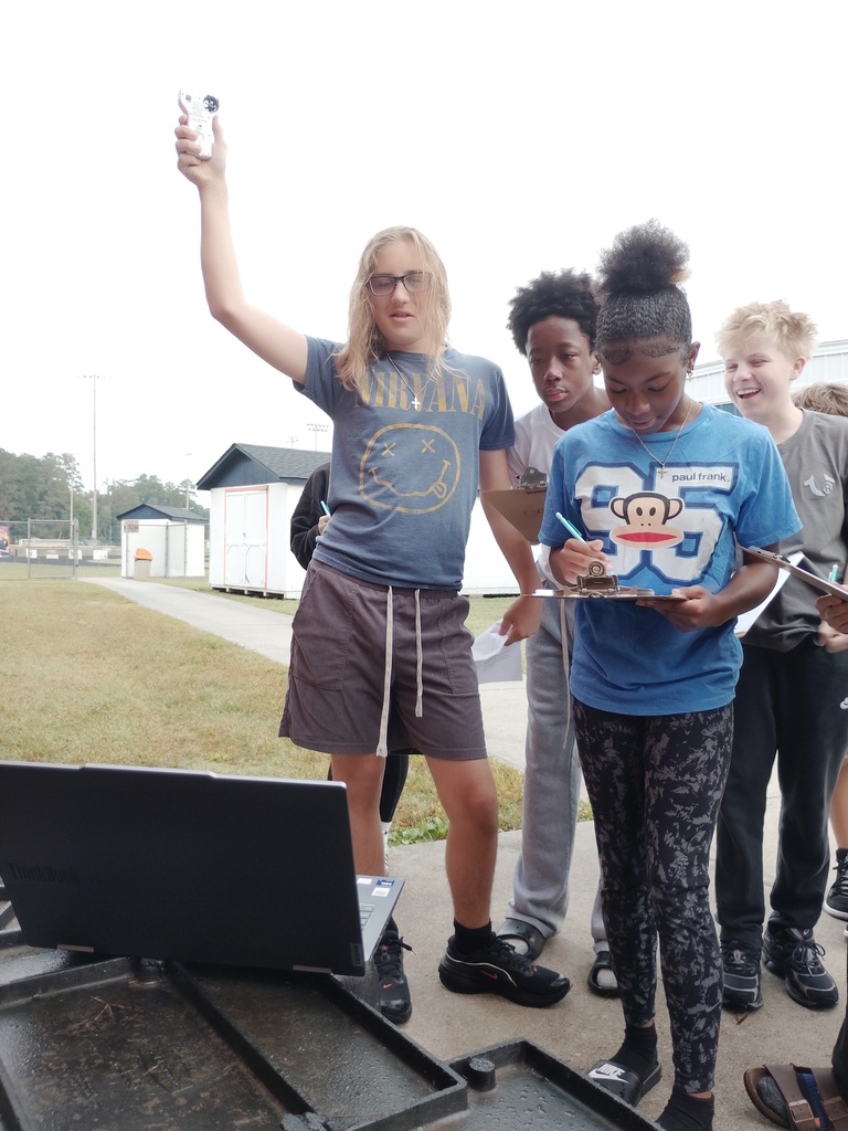 A group of students conduct an outdoor science experiment next to an open laptop. One student holds a handheld sensor up in the air while others stand nearby writing notes on clipboards.