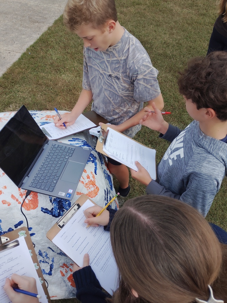 Three students gather around an outdoor painted rock with a laptop resting on top to complete work on clipboards. One student writes on a post-lab synthesis worksheet, another holds a clipboard while speaking, and a third takes notes in the background.