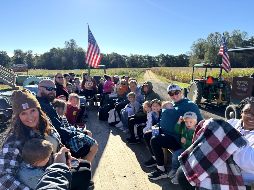 A group of children, parents, and teachers riding together on an outdoor wooden wagon hayride on a bright, clear day