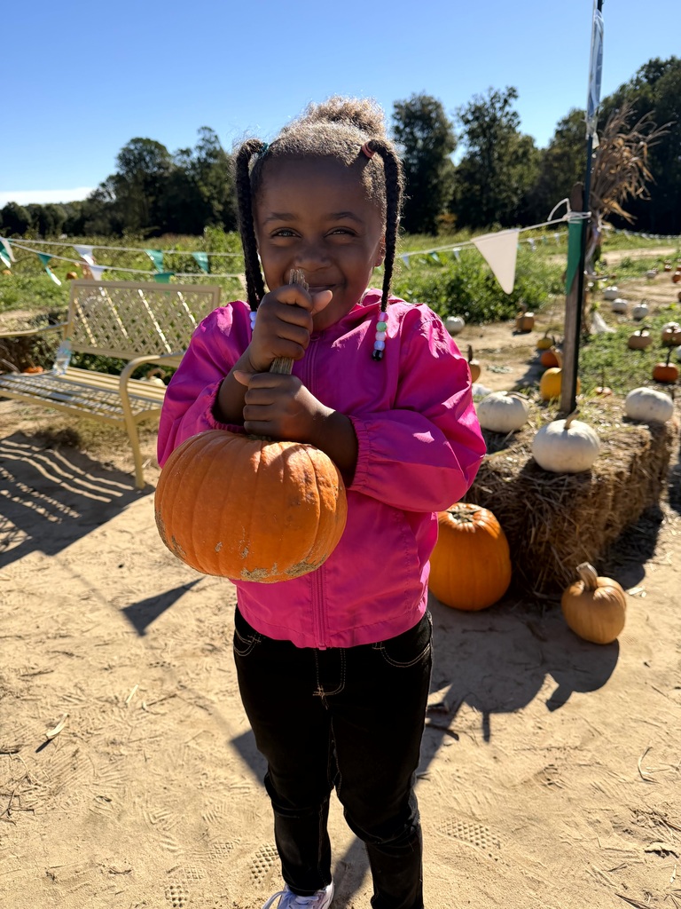 A smiling young girl wearing a bright pink jacket and holding up a medium-sized orange pumpkin in a pumpkin field.