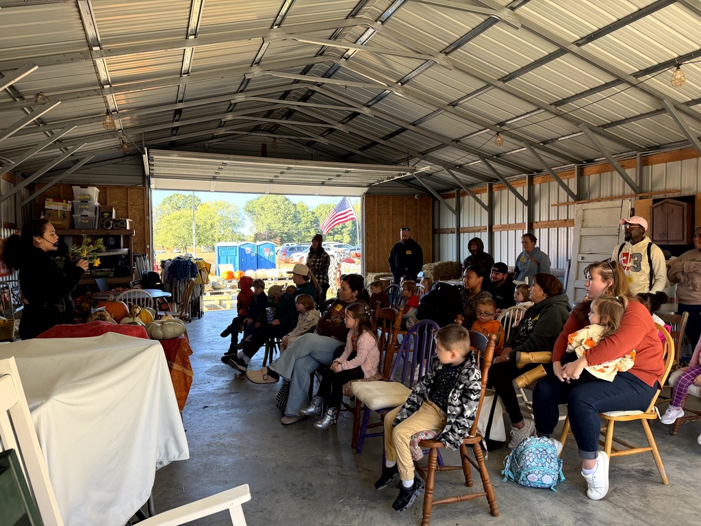 A group of children and adult chaperones sitting on chairs inside a large metal barn listening to a presenter at the front.